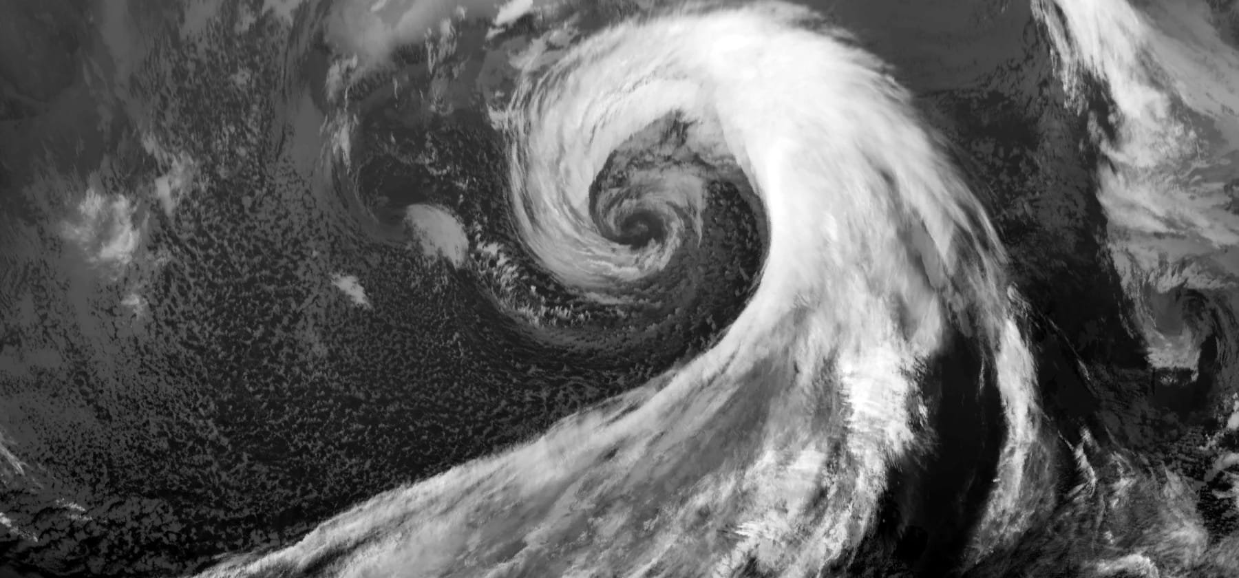 The same low in infrared at the same moment. The outer comma and the frontal band shine bright white because their tops are high and cold. The inner spiral is mid grey with a dark core because its tops are lower, and the shower cells that were bright in the visible image are dim, because their tops are low and not much colder than the sea.