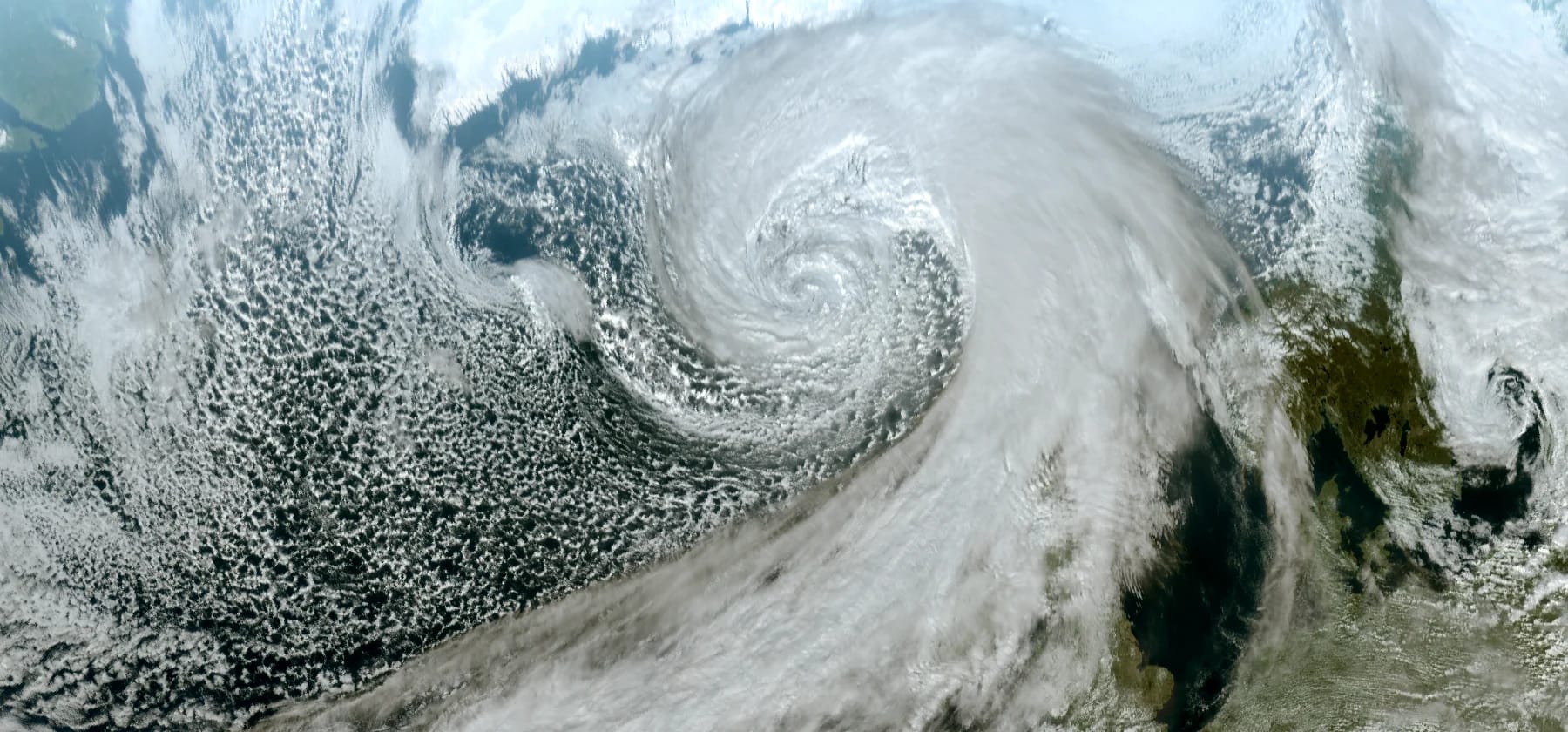 Real visible satellite image of a deep low south of Iceland. A huge comma of cloud has a tight spiral in its head and a long band sweeping away to the lower left, with a sharp edge on its cold-air side. A broad shield of cloud lies ahead of it to the upper right, and a honeycomb of small shower cells fills the cold air behind the band.